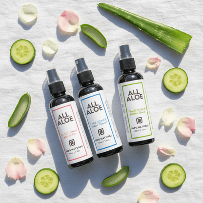 Three small spray bottles of face toner arranged on a white fabric background with sliced cucumbers, aloe vera slices, and pink flower petals. The bottles are labeled prasadhak ALL ALOE, with one pink label, one blue label, and one green label.