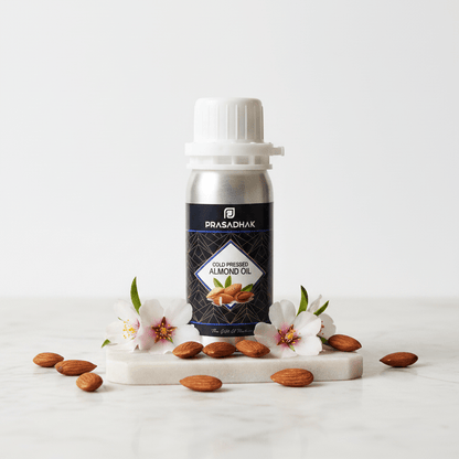 A bottle of Prasadhak Cold pressed Almond Oil stands on a white marble slab with scattered almonds and pink blossoms on a clean white background.