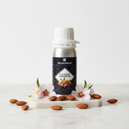 A bottle of Prasadhak Cold pressed Almond Oil stands on a white marble slab with scattered almonds and pink blossoms on a clean white background.