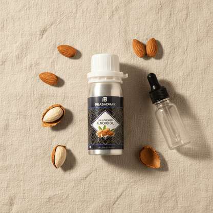 A bottle of Prasadhak Cold-Pressed Almond Oil is displayed on a textured beige linen background, surrounded by whole and halved almonds. A small, clear glass dropper bottle with a black cap lies to the right of the main product bottle.
