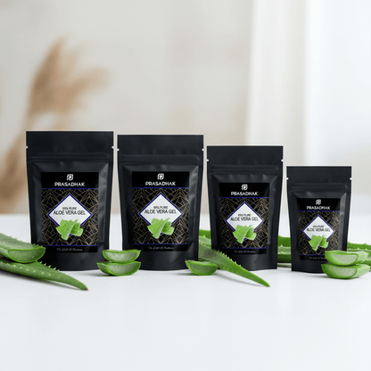 Four black stand-up pouches of Prasadhak Aloe Vera products arranged on a white surface with sliced aloe vera leaves around them. The pouches are labeled with the Prasadhak brand and different aloe vera gel/oil variants.
