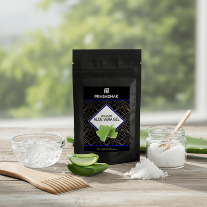 A black stand-up pouch of Prasadhak Aloe Vera Gel sits on a wooden surface with a blurred green outdoor background. Surrounding the pouch are a glass bowl of gel, aloe vera slices, a small jar of white powder with a wooden spoon, and a wooden comb.