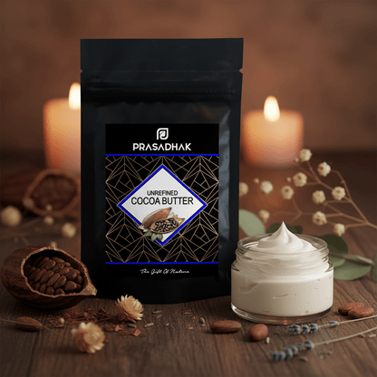 A stand-up pouch of Prasadhak Unrefined Cocoa Butter is displayed on a wooden surface next to a small glass jar filled with white cocoa butter cream, surrounded by almonds, dried flowers, and lit candles in the background.