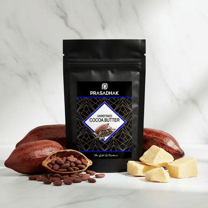 A black stand-up pouch of Prasadhak Unrefined Cocoa Butter stands on a white marble surface with cacao pods, a small bowl of cocoa beans, and pale yellow cocoa butter chunks arranged around it.