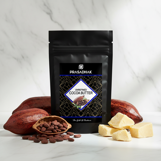 A black stand-up pouch of Prasadhak Unrefined Cocoa Butter stands on a white marble surface with cacao pods, a small bowl of cocoa beans, and pale yellow cocoa butter chunks arranged around it.