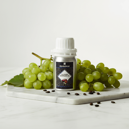 A bottle of Prasadhak Grape Seed Oil centered on a white marble slab, surrounded by green grapes, seeds, and leaves against a clean white background.