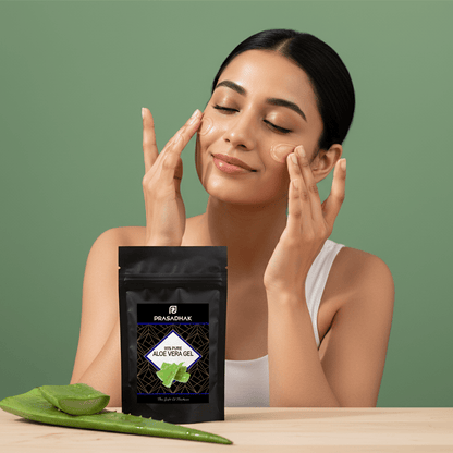 A woman with closed eyes applies aloe vera gel to her face, with a black stand-up pouch of PRABADHARX Aloe Vera Gel placed on a wooden table in the foreground, alongside fresh aloe vera leaves, against a solid sage green background.