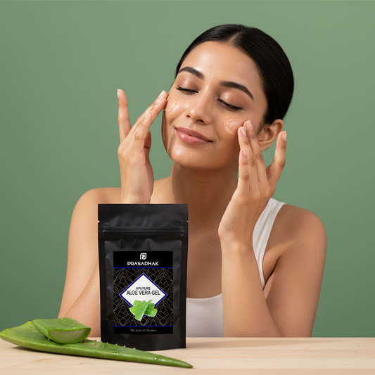 A woman with closed eyes applies aloe vera gel to her face, with a black stand-up pouch of PRABADHARX Aloe Vera Gel placed on a wooden table in the foreground, alongside fresh aloe vera leaves, against a solid sage green background.