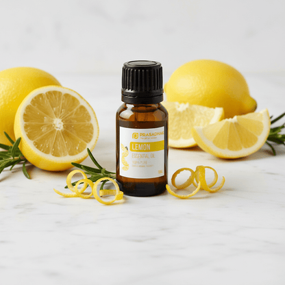 A bottle of Prasadhak Lemon Essential Oil centered on a light marble surface, surrounded by whole and sliced lemons, rosemary sprigs, and yellow citrus curls.