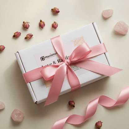 A white gift box wrapped with a wide pink satin ribbon and bow, placed on a light beige surface with the box brand Prasadhak.
