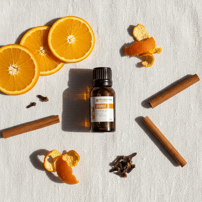 A small amber glass bottle labeled Prasadhak ORANGE essential oil with a black cap sits on a light fabric surface, surrounded by orange slices, orange peels, cinnamon sticks, and cloves in a styled flat-lay arrangement.