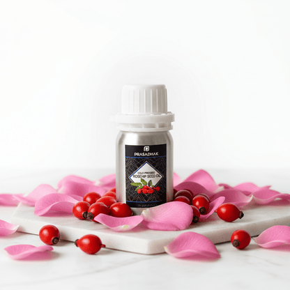 A small bottle of Prasadhak Rosehip Oil stands on a white marble slab surrounded by pink rose petals and red berries, against a clean white background.