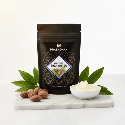 A black stand-up pouch of Prasadhak Unrefined Shea Butter stands on a white marble board with green leaves, nuts, and a small white bowl of pale cream-colored shea butter against a clean white background.