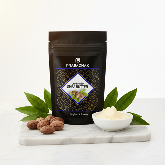 A black stand-up pouch of Prasadhak Unrefined Shea Butter stands on a white marble board with green leaves, nuts, and a small white bowl of pale cream-colored shea butter against a clean white background.