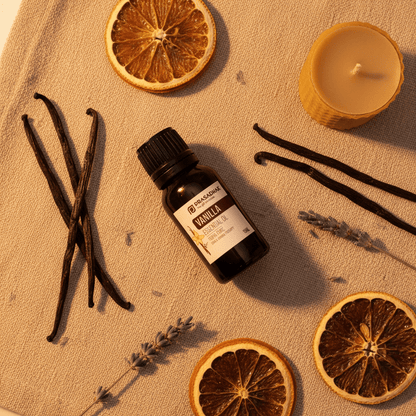 A bottle of Prasadhak Vanilla Oil with a black cap and white label, styled on a beige linen surface with dried orange slices, vanilla beans, lavender sprigs, and a lit candle.